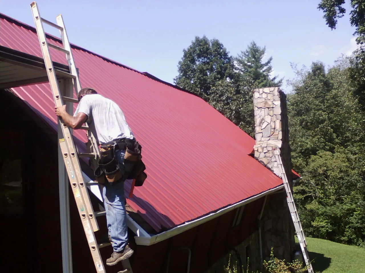 Roofer on a ladder beside a red metal roof and stone chimney — Soffit & Fascia Repair by Roofing Brothers in Hugo